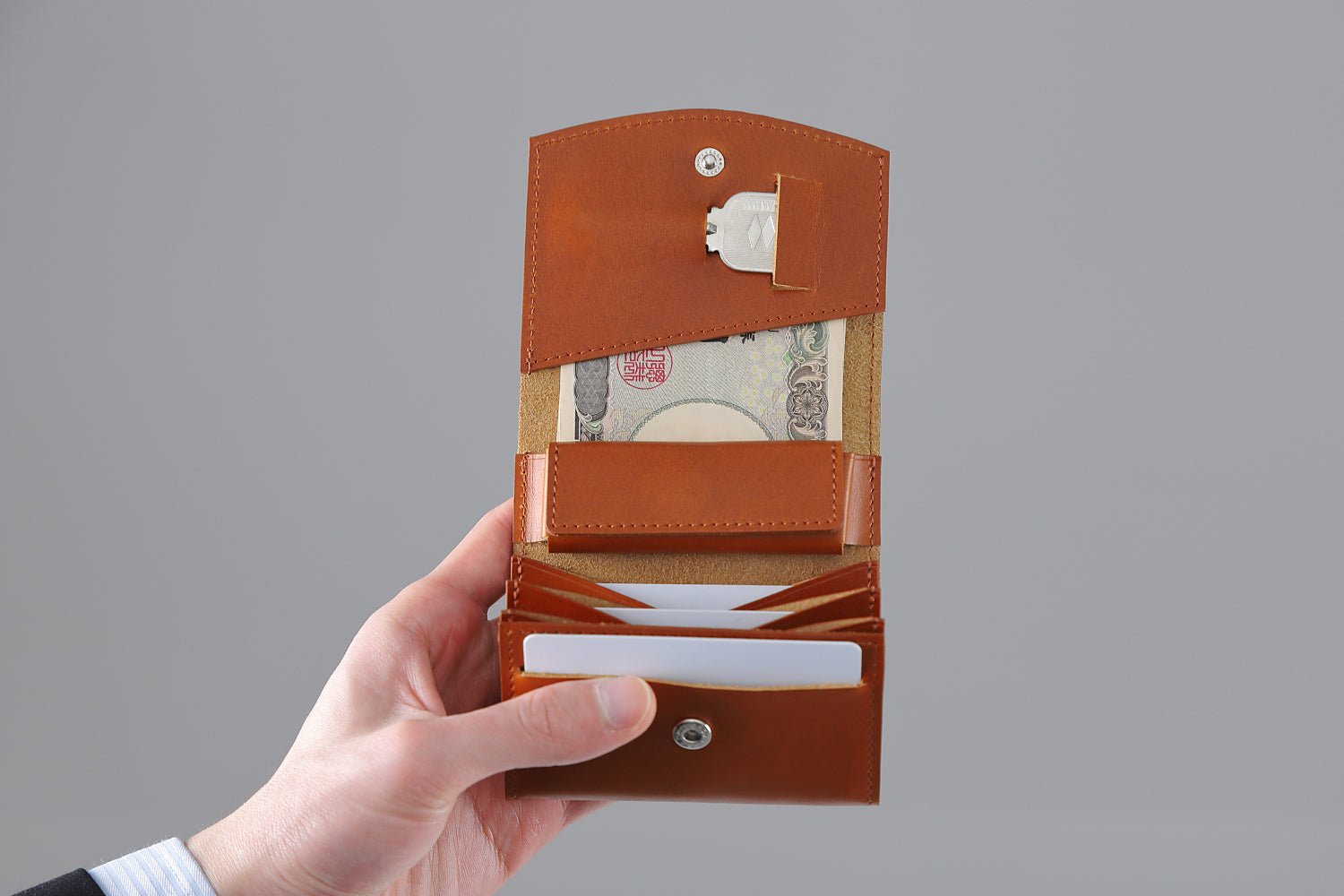 MC-bifoldwallet。蔵前の革職人が製作した国産牛革の薄いコンパクト二つ折り財布。キャメル。紙幣、カード、鍵を収納し、手で財布を見開いているシーン。