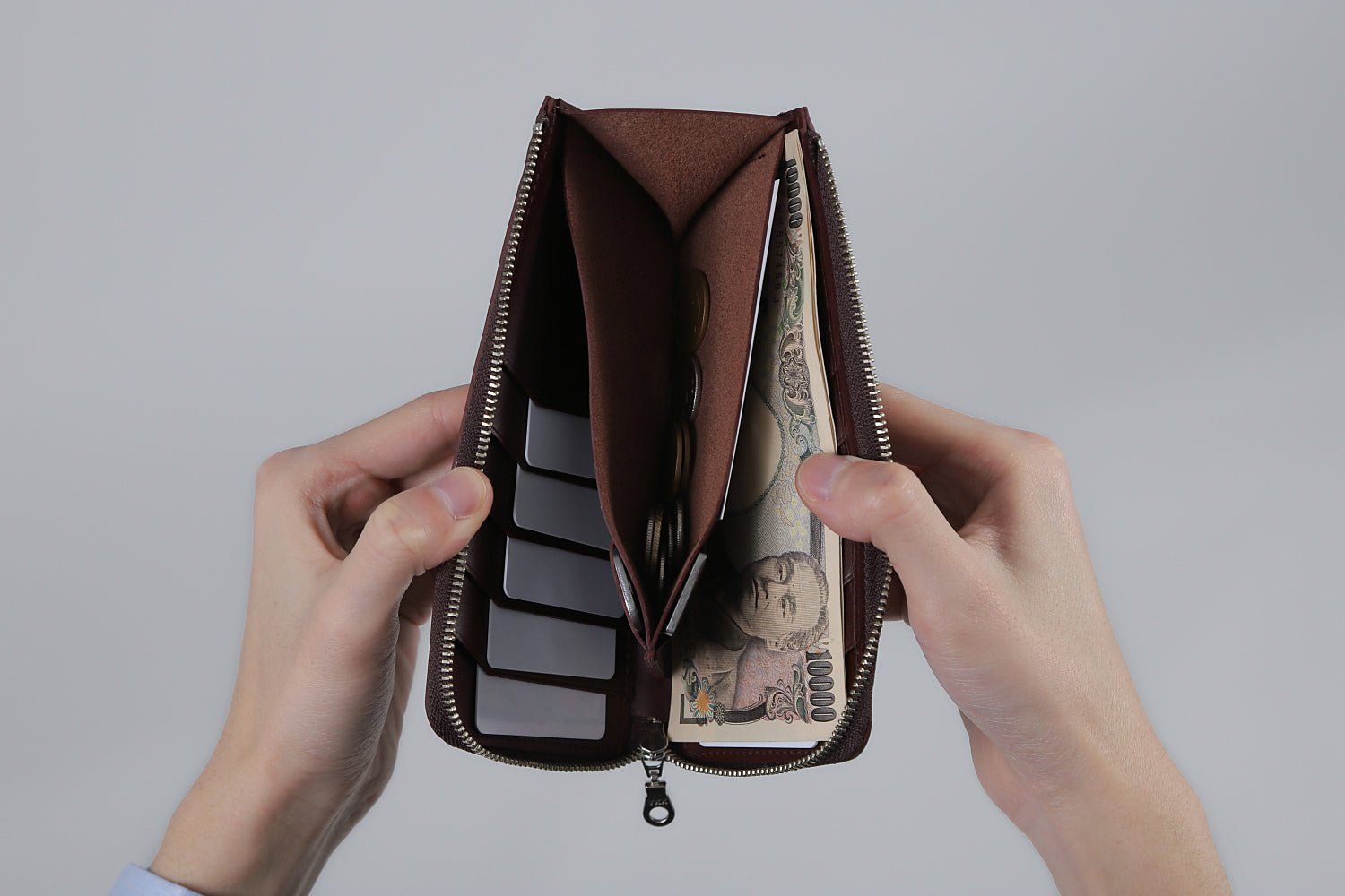 MC-longwallet。蔵前の革職人が製作した国産牛革のL字ファスナーコンパクト長財布。バーガンディ。紙幣、小銭、カード、鍵を収納し、手で財布を見開いているシーン。