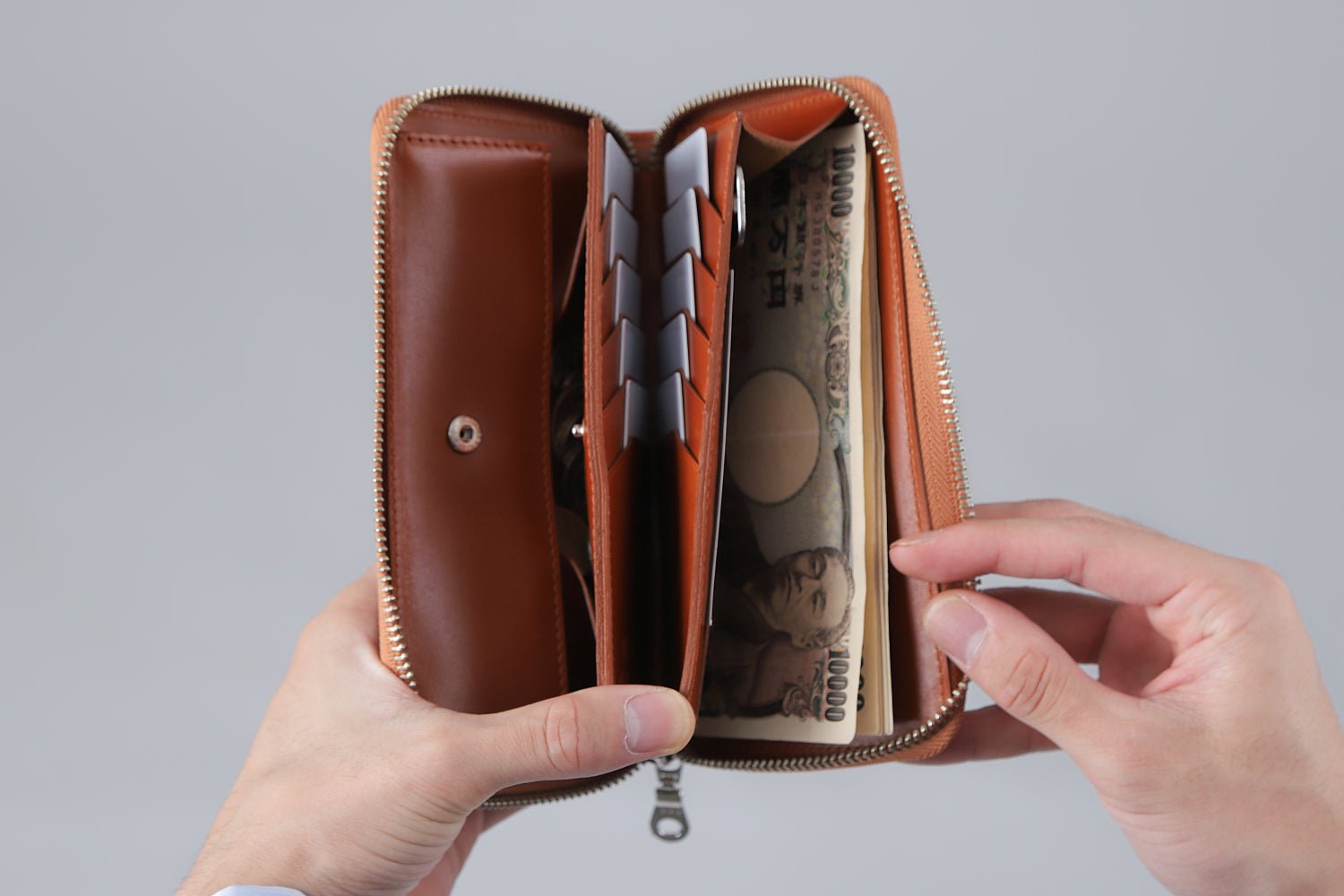 MC-longwallet2。蔵前の革職人が製作した国産牛革のラウンドファスナーコンパクト長財布。キャメル。紙幣、カード、鍵、小銭の収納し、手で財布を見開いているシーン。