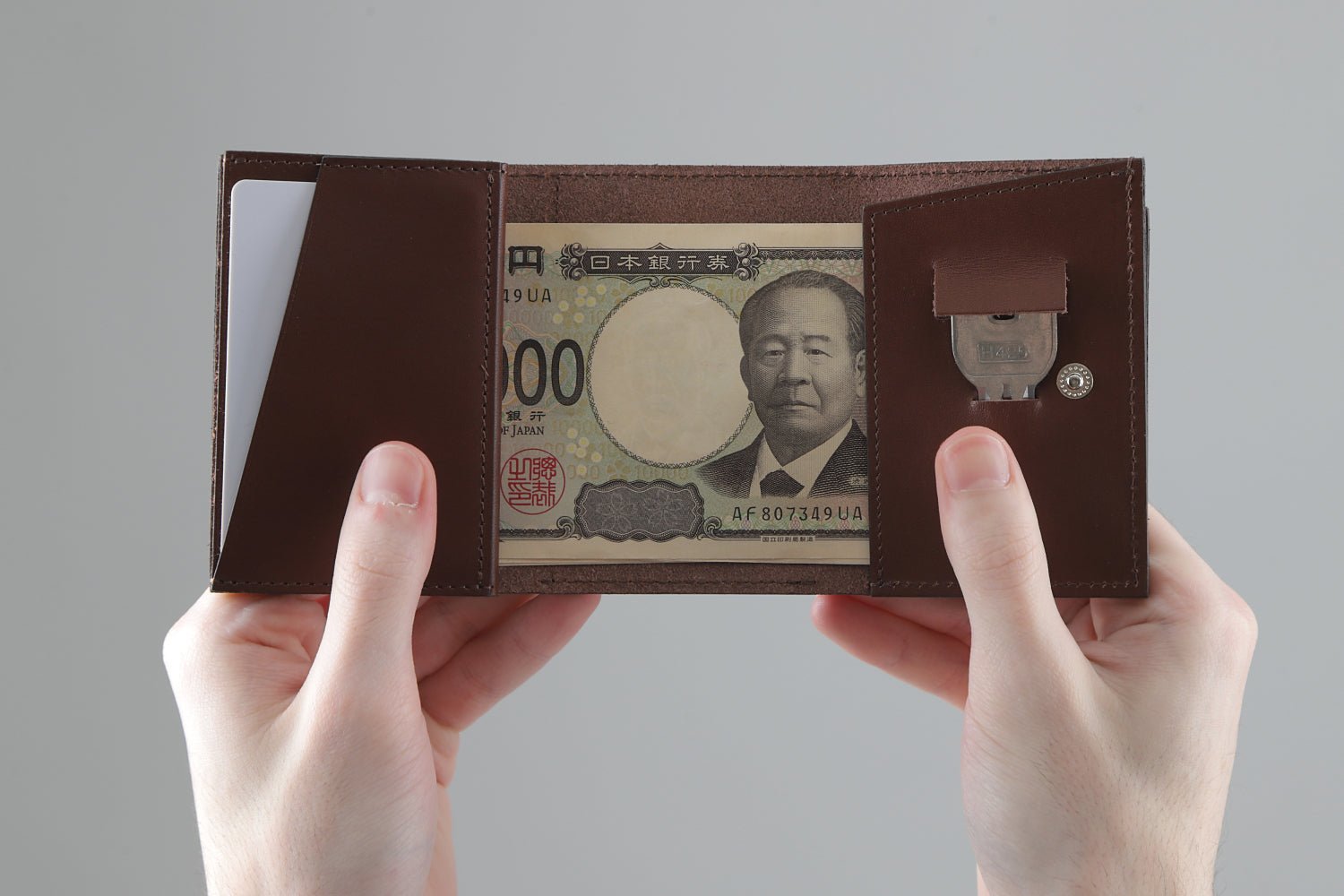 MC-miniwallet。蔵前の革職人が製作した国産牛革のミニ財布。ブラウン。カード、鍵、紙幣を収納し、手で財布を見開いているシーン。